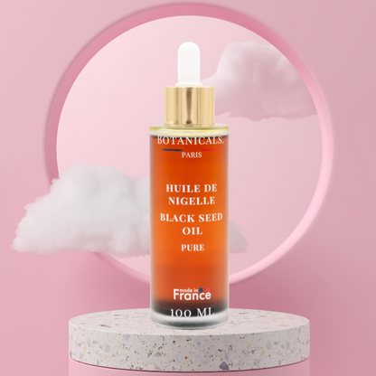 Une bouteille de 100ml d'huile de nigelle bio devant un fond rose, parsemé de léger nuage